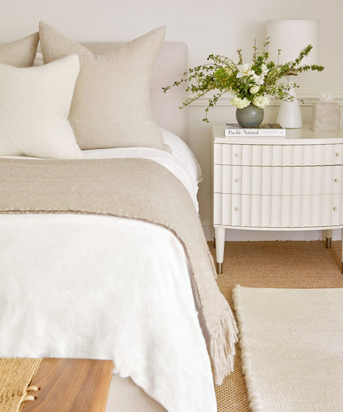 A cozy, neutral-toned bedroom features a bed topped with beige and white pillows and the Large Alpaca Basketweave Throw. A white nightstand holds a vase of flowers, lamp, book, and tissue box. A woven rug lies on the floor.