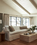 A bright, cozy living room with a Miramar Sofa, neutral cushions, a stone coffee table with greenery, large windows with beige curtains, and a woven rug under exposed white ceiling beams.