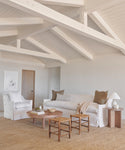 A bright, minimalist living room with white walls and exposed beams showcases a plush Miramar Sofa, an armchair, two wooden stools, a round coffee table on a jute rug, neutral cushions, and a table lamp.