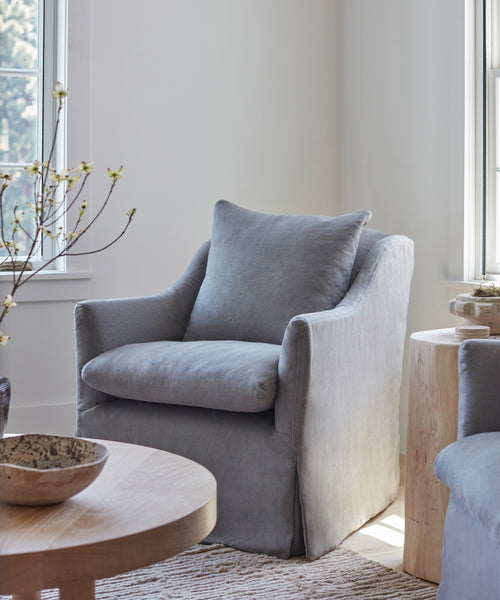 A cozy Miramar Chair Slipcover in light gray Belgian linen covers an armchair in a bright room near a window, paired with a matching cushion, round wood coffee table, pale wood side table, and set on a textured beige rug.