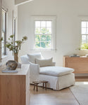 A bright, minimalist living room with large windows features the Miramar Chaise Slipcover in white linen, accented by pillows, light wood furniture, a small stool, and a dresser topped with a vase of greenery as sunlight pours in.
