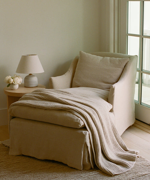 A cozy beige Miramar Chaise Slipcover drapes an armchair beside a round side table with a lamp and vase of white flowers, all set near a window with glass panes.
