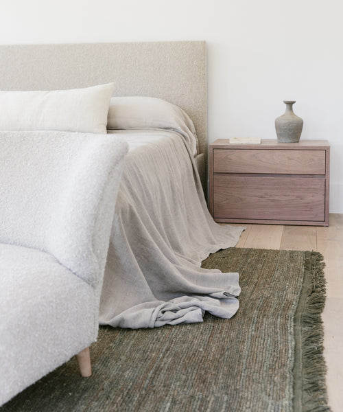 Minimalist bedroom with a beige upholstered bed, light bedding draped onto a green Mojave Jute Rug, a wooden nightstand featuring a ceramic vase and book, and a light-colored armchair in the foreground.