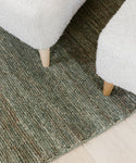 Close-up view of the corner of the Mojave Jute Rug, featuring a textured green design on a light wood floor, with two light beige upholstered furniture legs resting on the sustainable rug.