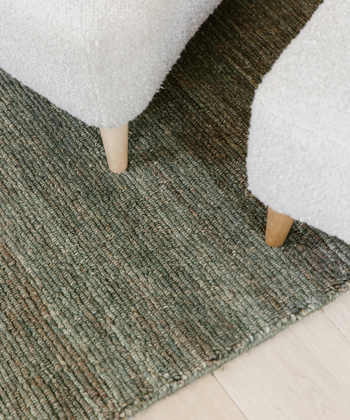 Close-up view of the corner of the Mojave Jute Rug, featuring a textured green design on a light wood floor, with two light beige upholstered furniture legs resting on the sustainable rug.