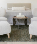 A cozy bedroom features a neutral bed, a rustic wooden side table with books and a candle, and two textured white armchairs, all set atop the Mojave Jute Rug for warmth and character.