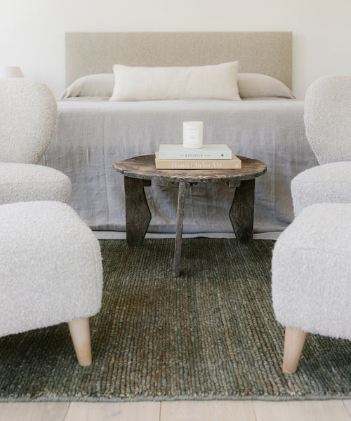 A cozy bedroom features a neutral bed, a rustic wooden side table with books and a candle, and two textured white armchairs, all set atop the Mojave Jute Rug for warmth and character.
