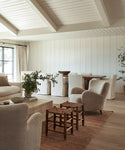 A bright, airy living and dining room with white paneled walls, cream furniture, wooden stools, green plants, and a large Mojave Jute Rug handmade in India creates a cozy, minimalist modern atmosphere.