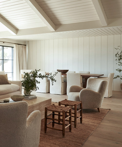 A bright, airy living and dining room with white paneled walls, cream furniture, wooden stools, green plants, and a large Mojave Jute Rug handmade in India creates a cozy, minimalist modern atmosphere.