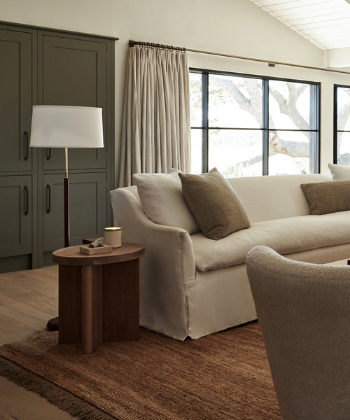 A cozy living room features a white sofa, brown cushions, wooden side table with candle, neutral curtains, large windows, a cabinet with muted green doors, and a handmade Mojave Jute Rug from India covering the wooden floor.