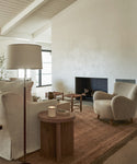 A cozy living room with neutral tones features a white armchair, a textured beige armchair, a round wooden side table with a lit candle, a floor lamp, and the Mojave Jute Rug handmade in India beside the modern fireplace.