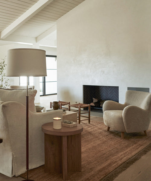 A cozy living room with neutral tones features a white armchair, a textured beige armchair, a round wooden side table with a lit candle, a floor lamp, and the Mojave Jute Rug handmade in India beside the modern fireplace.