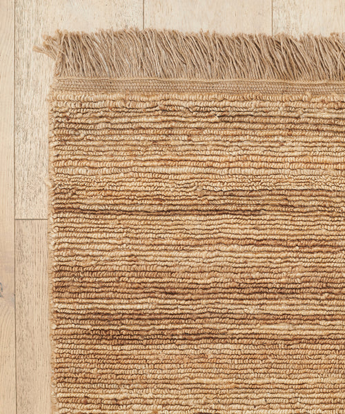 A close-up of the Mojave Jute Rug, handmade in India with horizontal ridges and fringed edges, shown on a light wooden floor.