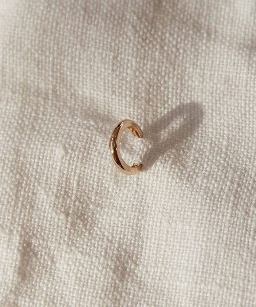 The Huggie, a small rose gold hoop earring, sits on textured off-white fabric, casting a soft shadow—a refined piece of minimalist jewelry.