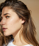 A young woman with long brown hair and fair skin is shown in close-up profile, wearing a white shirt and elegant Huggie with Diamonds earrings, as soft natural light highlights her face.