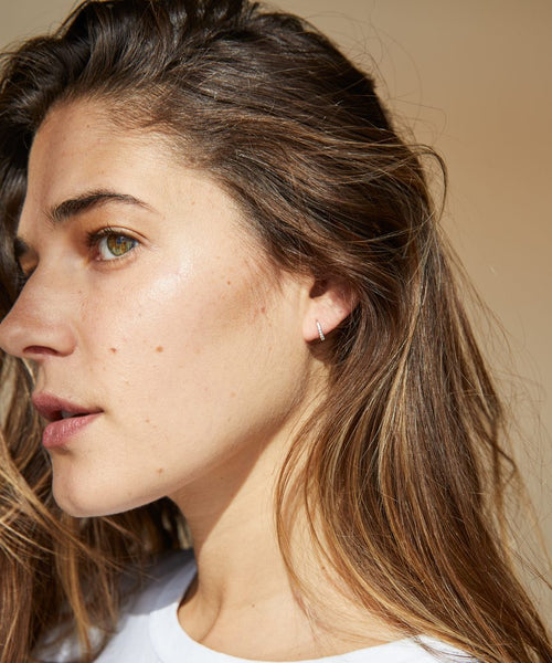 A young woman with long brown hair and fair skin is shown in close-up profile, wearing a white shirt and elegant Huggie with Diamonds earrings, as soft natural light highlights her face.