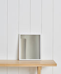 A square mirror with a white frame sits on a light wooden shelf against a white paneled wall, styled alongside the Untitled 5 Framed Print for an elevated Jenni Kayne Home aesthetic.