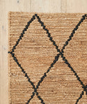 A close-up of the Wilder Diamond Jute Rug shows its bold black diamond pattern on a light wooden floor, with only the corner of the rectangular handwoven rug visible.