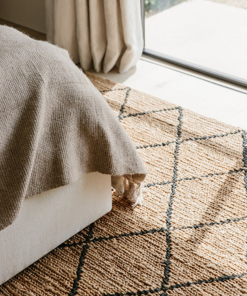 A cozy beige bed with a textured blanket sits next to the Wilder Diamond Jute Rug, a handwoven piece featuring a black diamond pattern. Sunlight streams through light curtains, creating a warm and inviting bedroom atmosphere.