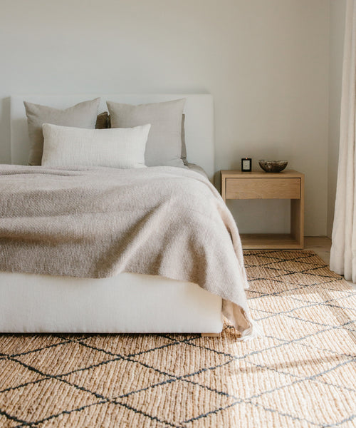 A cozy, minimalist bedroom features a white bed with beige bedding, neutral pillows, a wooden nightstand with a bowl and candle, and the Wilder Diamond Jute Rug on the floor as sunlight streams through the curtains.