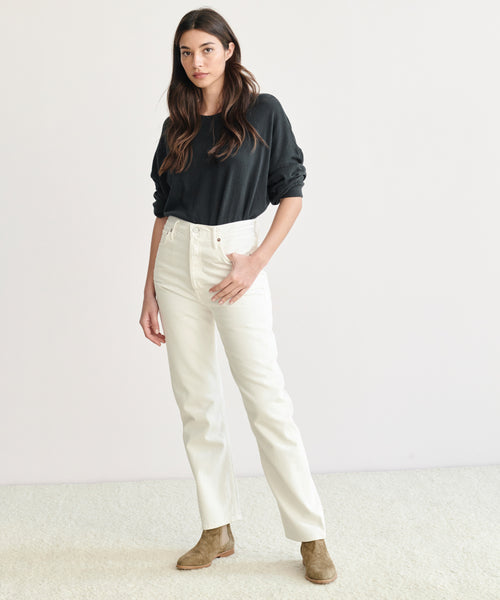 A woman with long brown hair stands against a plain background, wearing a black long-sleeve shirt tucked into 90s Pinch Waist Jeans and brown ankle boots.