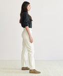 A woman with long dark hair stands in profile against a plain background, wearing a black long-sleeve top, 90s Pinch Waist Jean in white, and brown boots.