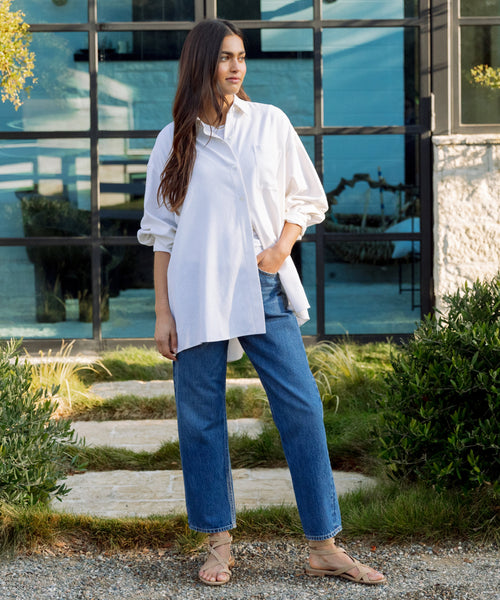 A person stands outdoors on a stone path, wearing a loose white button-up shirt and the 90s Crop Jean in vintage denim blue with sandals. Green bushes line the path, set against a modern glass building in the background.