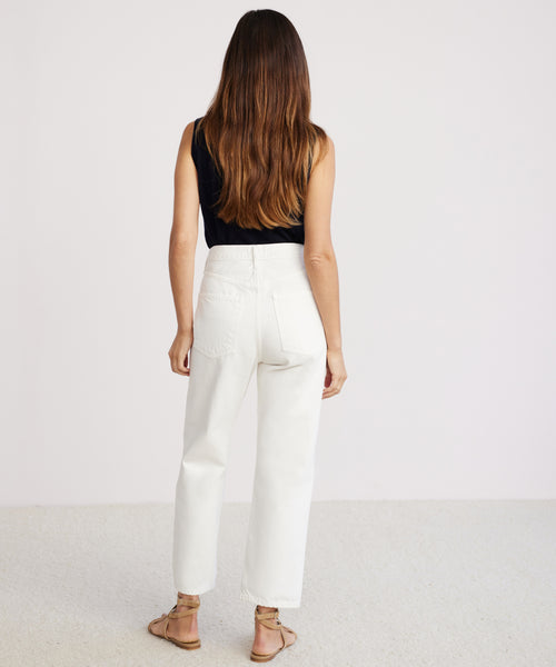 A woman with long brown hair, wearing a black sleeveless top, 90s Crop Jean in a white vintage style, and sandals stands facing away from the camera against a light-colored background.