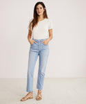 A woman stands against a plain white background, wearing the Riley Crop Jean in light blue stretch denim with a white short-sleeve T-shirt and tan sandals. Her hands are in her pockets as she looks at the camera with a neutral expression.