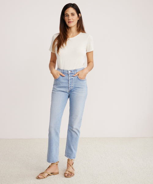 A woman stands against a plain white background, wearing the Riley Crop Jean in light blue stretch denim with a white short-sleeve T-shirt and tan sandals. Her hands are in her pockets as she looks at the camera with a neutral expression.