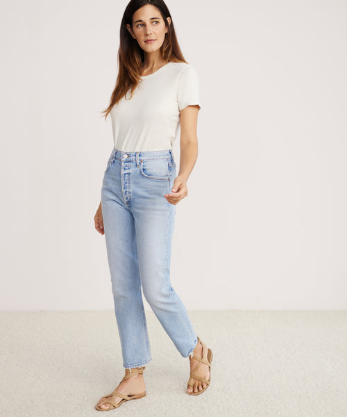 A woman stands indoors wearing a white short-sleeve shirt, Riley Crop Jean in light blue stretch denim, and tan sandals, looking slightly to the side with one hand near her waist.
