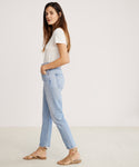 A woman with long brown hair stands sideways against a plain light background, wearing a white organic cotton t-shirt, tan sandals, and Riley Crop Jean in light blue, with her hands in her pockets.