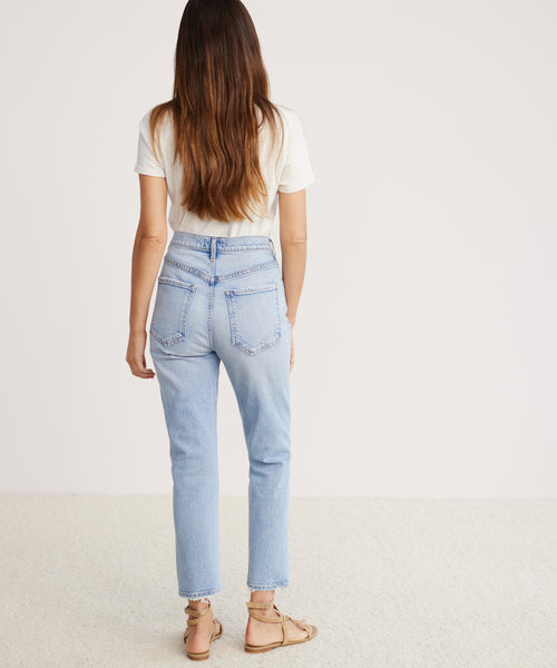 A woman with long brown hair, wearing a white t-shirt and Riley Crop Jean in light blue denim with tan sandals, stands facing away from the camera in a plain, light-colored room.