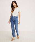 A woman in a white t-shirt and the Riley Crop Jean stands on a light floor against a pale background, wearing beige sandals and looking down with a relaxed expression.