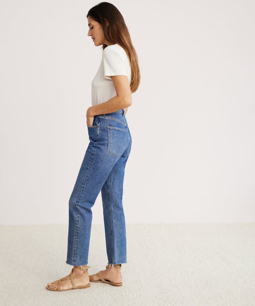 A woman with long brown hair stands sideways on a light carpet, wearing tan sandals, a white short-sleeve tee, and blue Riley Crop Jeans made from stretch denim, with one hand in her pocket.
