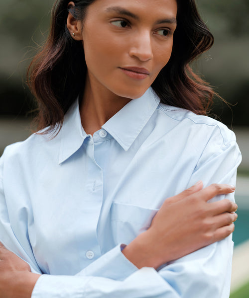 A woman with wavy brown hair wears the Boyfriend Shirt outdoors, crossing her arms and gazing thoughtfully to the side.