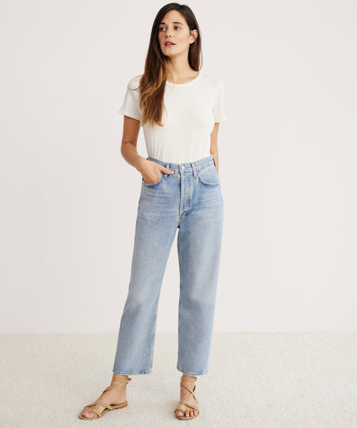 A woman with long brown hair stands against a plain background, wearing a white short-sleeve t-shirt, 90s Crop Jean in light blue organic cotton denim, and beige sandals, with one hand in her pocket.