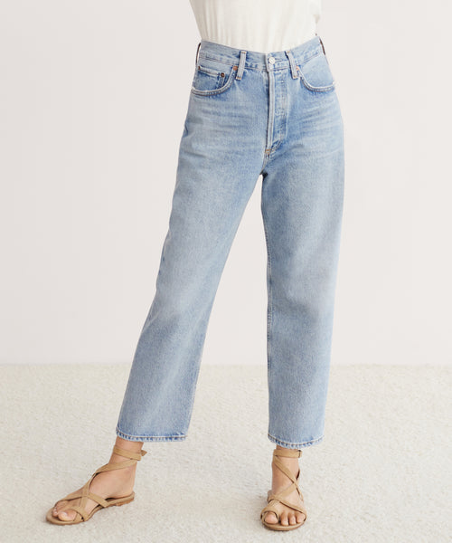 Person wearing AGOLDE 90s Crop Jean in light blue organic cotton denim, a white tucked-in top, and tan strappy sandals, standing against a plain light background. Only the lower body is visible.