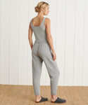 A woman stands on a woven rug facing away, wearing the light gray Saturday Sweatpant in ultra-soft French terry with dark gray slippers. She is in front of a white paneled wall, her blonde hair tied up.