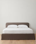 A minimalist bed with a brown upholstered base and headboard, dressed in crisp white sheets and pillows, features the Cove Bed Slipcover. It stands on a light wooden floor against a plain white wall.