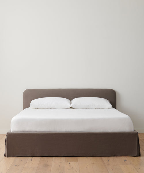 A minimalist bed with a brown upholstered base and headboard, dressed in crisp white sheets and pillows, features the Cove Bed Slipcover. It stands on a light wooden floor against a plain white wall.
