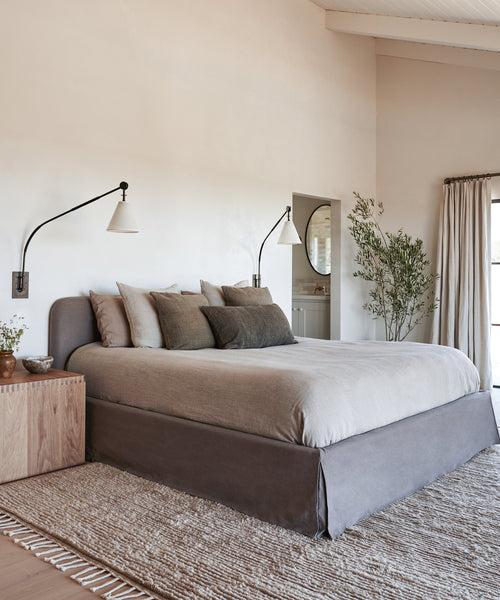 A modern bedroom with the Cove Bed, featuring an upholstered headboard and neutral bedding, throw pillows, a wooden nightstand, wall-mounted reading lamps, an indoor plant, a woven rug, and light walls lit by natural light.