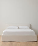 A neatly made bed features an ivory linen upholstered headboard, two pillows, a white bedspread, and the Cove Bed Slipcover by Belgian Libeco, set against a plain white wall with light wood flooring.