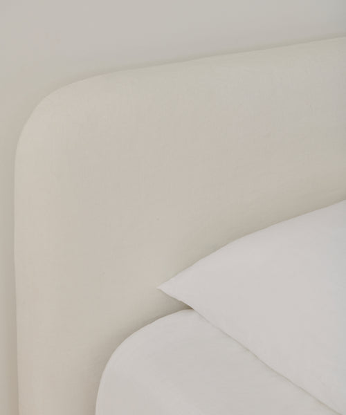 Close-up of the Cove Bed in ivory linen with a rounded, upholstered headboard and white Belgian Libeco linen bedding, highlighting its soft, minimalist design against a light wall.