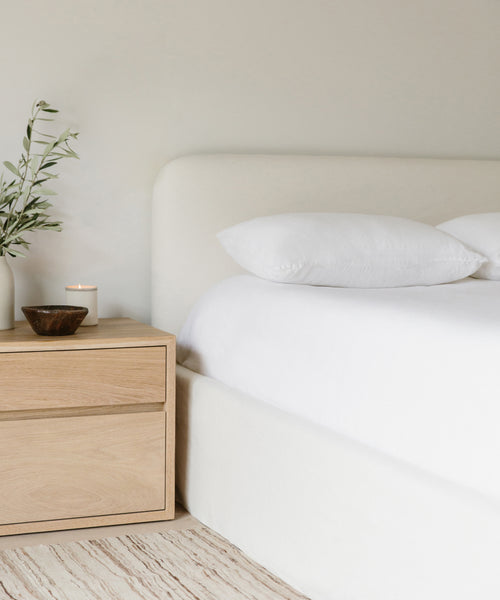 A minimalist bedroom highlights the Cove Bed in ivory linen with white pillows, set against a cream wall, and a light wood nightstand topped with a vase of green branches, a lit candle, and a small bowl.