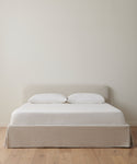 The Cove Bed Slipcover features a light beige upholstered headboard and base, styled with natural linen bedding and two white pillows, positioned against an off-white wall on a wooden floor for a minimalist look.