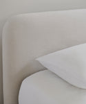 A close-up of the Cove Bed Slipcover in light beige linen features a headboard with white bedsheets and pillow, set against a light wall and highlighting its elegant natural texture.