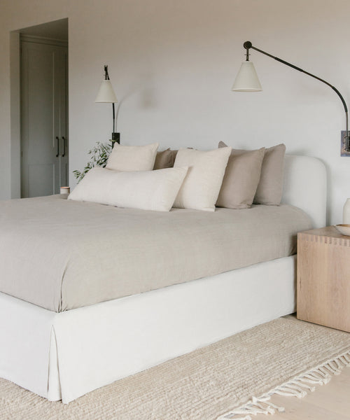 A minimalist bedroom features a neatly made Cove Bed Slipcover in natural linen, several pillows, a wooden nightstand, wall-mounted lamps, and a light woven rug for a calm, modern look.