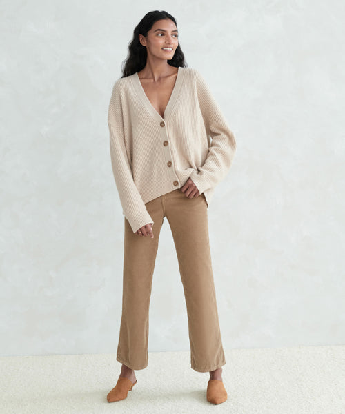 A woman wearing the Cropped Cashmere Cocoon Cardigan stands against a light gray textured wall, paired with tan wide-leg pants and brown slip-on shoes. She has long dark hair, smiles slightly, and poses with one hand in her pocket.