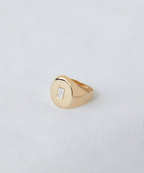 The Gemma Baguette Signet Ring showcases a rectangular baguette-cut diamond set in 14k gold on a light gray fabric background—a classic piece from Jenni Kayne Jewelry.